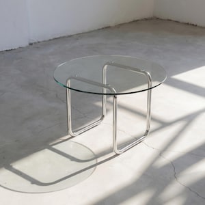 May include: A modern, round coffee table with a clear glass top and a polished chrome frame. The table's legs are a continuous, curved design. The table is approximately 18 inches in height and 30 inches in diameter.