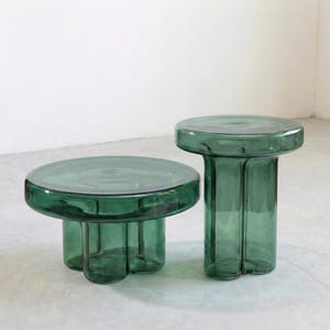May include: Two transparent green glass tables. One is round with a three-footed base, and the other is a taller, cylindrical table. Both have a smooth, reflective surface and are made of thick glass.