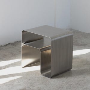 Op de afbeelding: Een bijzettafel van geborsteld metaal met een minimalistisch ontwerp. De tafel heeft een rechthoekige vorm met afgeronde hoeken en een onderste plank. Het metalen oppervlak reflecteert het licht. Een modern meubelstuk voor een eigentijds interieur.