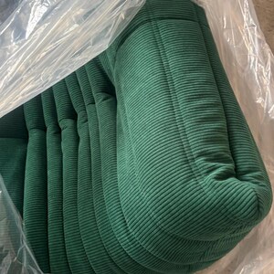 May include: A stack of green corduroy cushions, partially encased in clear plastic. The cushions have a ribbed texture and a rounded shape, suggesting they are for seating or decorative purposes.