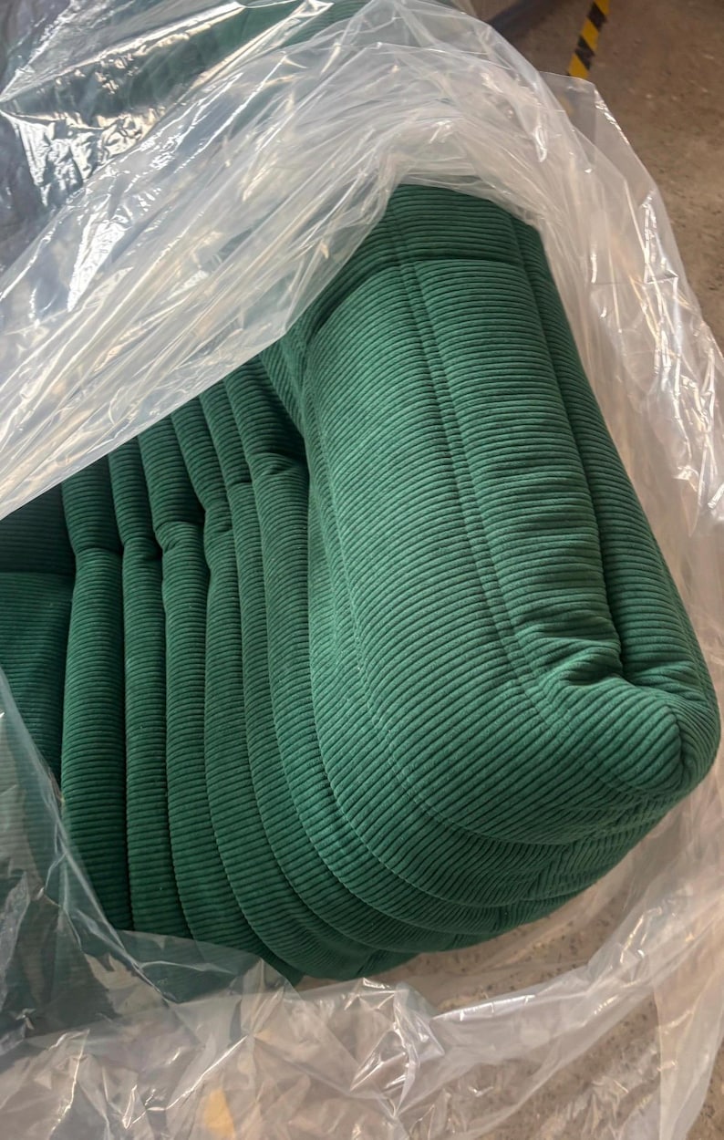 May include: A stack of green corduroy cushions, partially encased in clear plastic. The cushions have a ribbed texture and a rounded shape, suggesting they are for seating or decorative purposes.