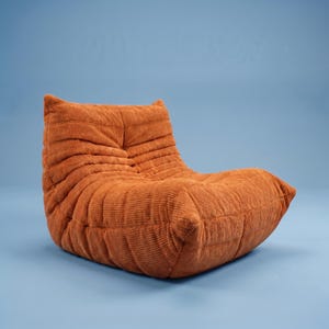 May include: An orange corduroy lounge chair with a low profile and a relaxed, inviting design. The chair has a rounded shape with a series of horizontal ridges, creating a textured appearance. The chair is set against a solid blue background.