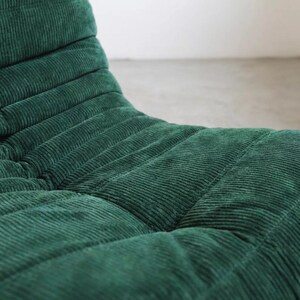 May include: A close-up of a green corduroy chair. The chair has a low profile and a curved, ergonomic design. The corduroy fabric is a deep shade of green, with a ribbed texture. The chair appears to be well-padded and comfortable.