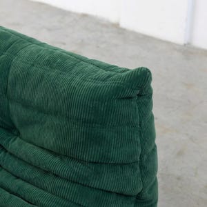 May include: Close-up of a section of a green corduroy sofa. The fabric has a ribbed texture and is a deep shade of green. The sofa appears to be well-padded and has a rounded shape. The design is modern and inviting.