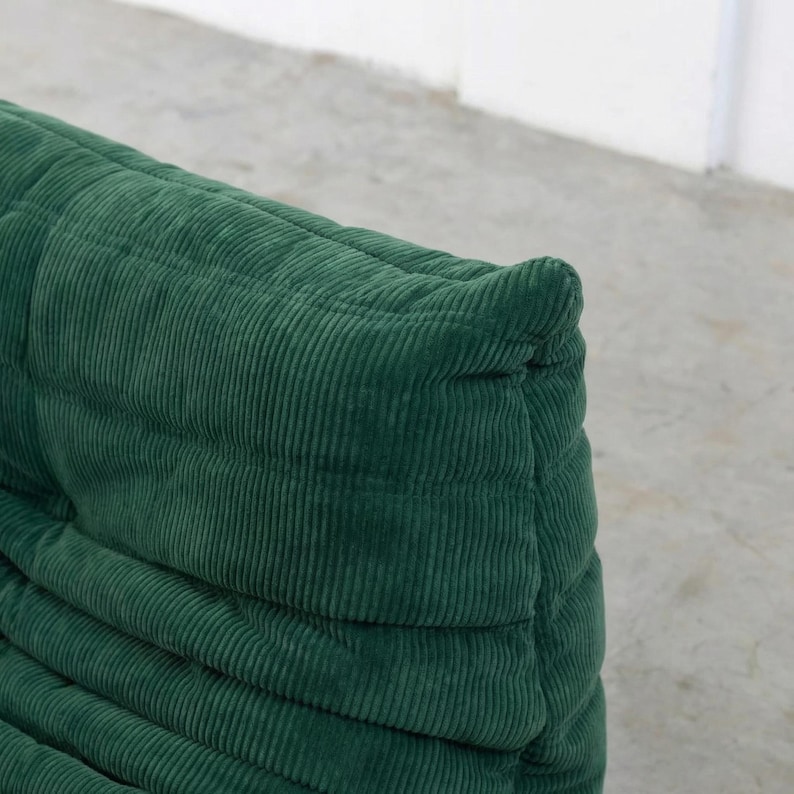 May include: Close-up of a section of a green corduroy sofa. The fabric has a ribbed texture and is a deep shade of green. The sofa appears to be well-padded and has a rounded shape. The design is modern and inviting.