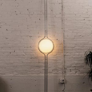 Puede incluir: Una lámpara de pie moderna y minimalista con una pantalla circular de vidrio esmerilado. La lámpara tiene una estructura delgada de metal plateado que se extiende desde la base hasta el techo. La luz está encendida, iluminando la habitación con un brillo suave. La lámpara está contra una pared de ladrillos blancos. Mide unos 200 cm.