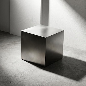 May include: A solid, metallic cube with a smooth, brushed finish. The object features sharp, clean edges and a uniform, reflective surface that catches light, creating subtle gradients across its flat, square faces and geometric form.