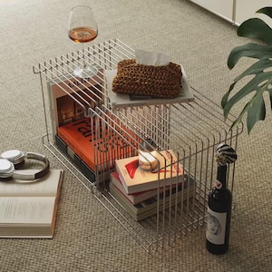 May include: A silver wireframe side table with books, a wine glass, and a bottle of wine. The table has two levels, with books and a woven tissue box on the top shelf. Headphones and an open book are on the floor.