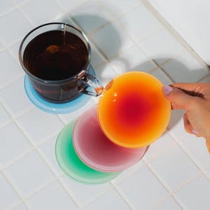 Op de afbeelding: Een glazen theekopje met thee staat op een lichtblauwe onderzetter. Daaronder liggen drie gekleurde onderzetters: groen, roze en oranje. Een oranje onderzetter wordt door een hand vastgehouden.