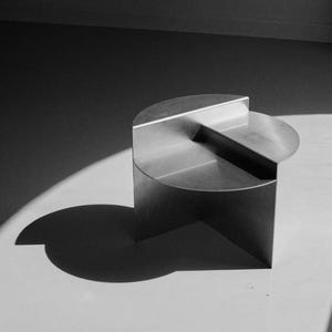 Puede incluir: Una mesa auxiliar moderna y minimalista en un entorno monocromático. La mesa está construida en metal cepillado, con una parte superior semicircular y una superficie inferior empotrada. El diseño incluye una base geométrica, proyectando una sombra.