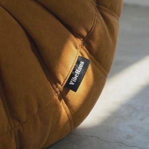May include: Close-up of a mustard-colored, quilted fabric. A black tag with the white text "VibeHaus" is sewn onto the fabric. The fabric has a soft, velvety texture and is stitched in a diamond pattern.