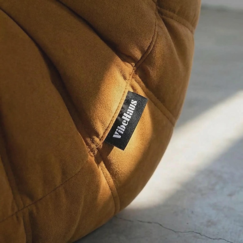 May include: Close-up of a mustard-colored, quilted fabric. A black tag with the white text "VibeHaus" is sewn onto the fabric. The fabric has a soft, velvety texture and is stitched in a diamond pattern.