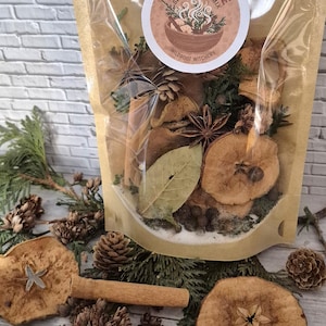 Może przedstawiać: Przezroczysta torebka celofanowa wypełniona mieszanką do potpourri z przyprawami jabłkowymi. Torebka zawiera suszone plasterki jabłek, laski cynamonu, anyż gwiazdkowaty, liście laurowe i inne przyprawy. Torebka ma okrągłą etykietę z napisem "Apple Spice Simmer Pot Mix" i "Wildroot Witchery".