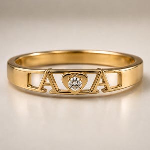 May include: A gold band ring featuring two capital letter A characters flanking a central heart-shaped setting. The heart contains a single round, clear, faceted gemstone at its centre. The ring has a polished, smooth, metallic finish throughout.