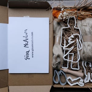 May include: An open cardboard box reveals a craft kit with various materials. Inside, there's a white card with the text "Gina McAdam" and a website address. The kit includes white plastic figure-shaped cutters, bundles of fiber, and a wooden rolling pin.