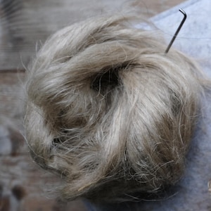 Puede incluir: Primer plano de una bola de fibra de lino natural, de color beige claro, con una herramienta metálica insertada. Las fibras de lino están dispuestas sueltas, mostrando textura y detalles. El fondo es borroso, de tono neutro.