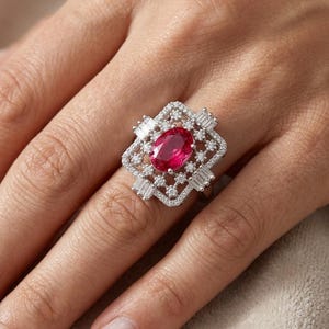 May include: A silver ring featuring a large, oval-cut ruby center stone. The ruby is surrounded by a geometric design of smaller, clear baguette and round-cut stones, creating a sparkling, elegant look. The ring is set on a silver band.