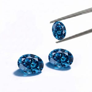 May include: Three oval-cut blue gemstones with a faceted surface that reflects light. One gemstone is held by a pair of metal tweezers, while the other two are positioned below it on a plain white surface, showcasing deep blue colour saturation.