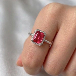 May include: A silver ring with a rectangular, bright red gemstone, encircled by a halo of small, clear stones. The ring sits on a slender band, highlighting the central stone's colour and sparkle. The ring is a piece of jewellery.