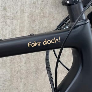 Op de afbeelding: Een zwart fietsframe met de tekst "Fahr doch!" in gouden letters. De fiets heeft een zwarte stuurpen en een zwarte band. Het frame is gemaakt van een matzwart materiaal.