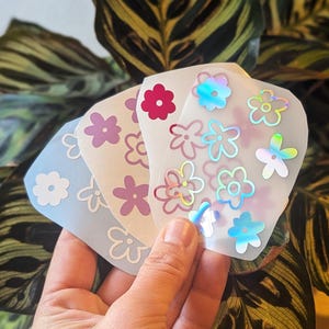 Op de afbeelding: Een verzameling van drie semi-transparante stickervellen met diverse bloemvormen. De vellen tonen bloemmotieven in effen wit, tinten paars, een helder rood en een reflecterende holografische afwerking die van kleur verandert in het licht.