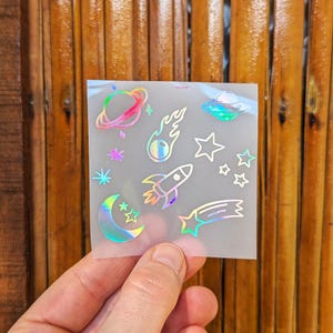Może przedstawiać: Arkusz holograficznych naklejek o tematyce kosmicznej z planetami, gwiazdami, rakietą, kometą i UFO. Naklejki mają tęczowe, opalizujące wykończenie. Drewniane tło.