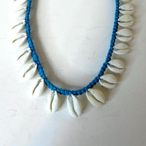 May include: A necklace featuring white seashells strung along a vibrant blue cord. The necklace has a braided design and adjustable blue ties. The seashells are evenly spaced around the necklace.