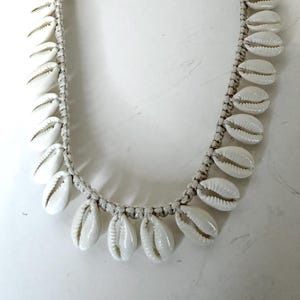 May include: A white cowrie shell necklace. The necklace features multiple white cowrie shells strung together with a woven, off-white cord. The shells are arranged in a symmetrical pattern, creating a beachy, bohemian aesthetic.