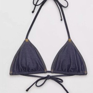 May include: A denim triangle bikini top with adjustable ties at the neck and back. The dark blue fabric is accented with light brown stitching and small gold-colored rivets at the top corners of the cups. A classic swimwear piece.