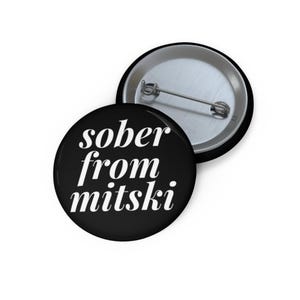 May include: A round black pin-back button featuring the white text sober from mitski in a serif font. The button is shown from two angles, displaying the front face with the text and the back side with a silver metal pin mechanism for attachment.