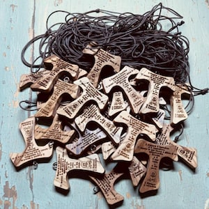 Può includere: Un gruppo di croci in legno con cordini grigio scuro. Ogni croce presenta un testo inciso e una piccola immagine. Le croci sono marroni chiare e sono disposte su una superficie azzurra anticata.