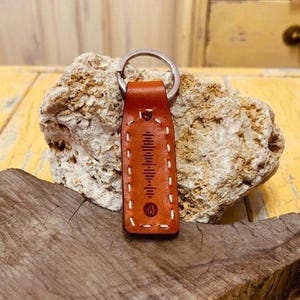 Può includere: Un portachiavi in ​​pelle marrone con un anello argentato. Il portachiavi rettangolare presenta un design cucito e un motivo a onde sonore. Il portachiavi è esposto su un pezzo di legno e una roccia.