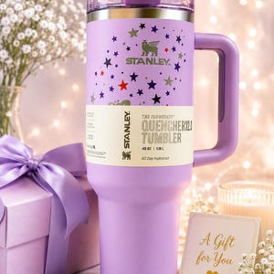 May include: A lavender Stanley Quencher H2.0 FlowState tumbler with a 40 oz capacity. The cup features a pattern of small multi-colored stars and a handle. A paper band around the center displays the text Stanley The Flowstate Quencher H2.0 Tumbler.