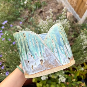 May include: A cylindrical ceramic planter featuring a textured, unglazed tan base and a reactive glaze finish in shades of light blue, turquoise, and white. The upper rim is shaped with two distinct, pointed peaks, creating a V-shaped cutout.
