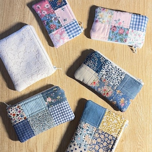Può includere: Una collezione di pochette con cerniera patchwork fatte a mano in varie dimensioni e fantasie. Le pochette presentano un mix di tessuti floreali, a pois e a quadri nei toni del blu, rosa e giallo. Una pochette è bianca con un disegno in pizzo.
