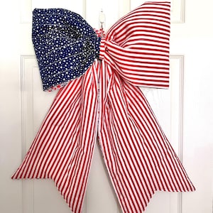 May include: A large patriotic bow with a blue fabric section covered in white stars and a red and white striped section. The bow is designed to be hung on a door or wall, perfect for Independence Day or other patriotic holidays.
