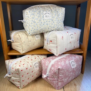 May include: Five quilted cosmetic bags in various floral patterns and colors, including blue, cream, and pink. Each bag features a white zipper and a small label with text. The bags are displayed on a wooden shelf.