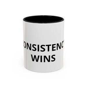 Peut inclure: Mug en céramique blanche avec intérieur noir et l'inscription "CONSISTENCY WINS" en noir. La tasse est cylindrique et semble vide.