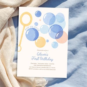 May include: A cream-colored invitation with watercolor bubbles in shades of blue, yellow, and peach. The text "POP ON OVER" is above the bubbles. The invitation reads "Oliver's First Birthday" with event details.