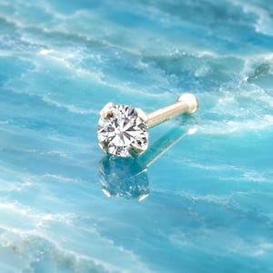 May include: A gold nose stud featuring a single round brilliant-cut clear gemstone held in a four-prong setting. The post is straight with a small rounded end, reflecting light clearly against the surface beneath the jewelry piece.