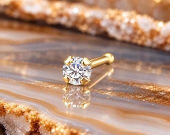 May include: A gold nose stud featuring a round, clear, faceted gemstone held in a four-prong setting. The post is straight with a small, rounded ball end, and the metal has a polished, reflective finish that catches the light.