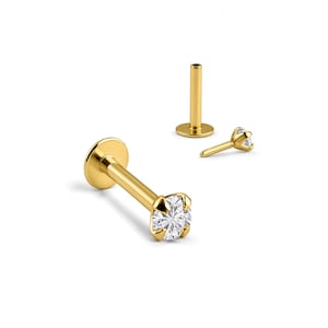 May include: A gold-toned labret stud earring featuring a clear, round-cut gemstone held in a four-prong setting. The piece includes a flat circular back disc and a straight post, shown alongside a separate matching post and a detached gemstone top.