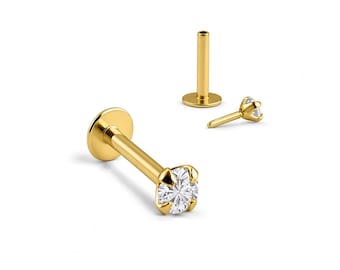 May include: A gold-toned labret stud earring featuring a clear, round-cut gemstone held in a four-prong setting. The piece includes a flat circular back disc and a straight post, shown alongside a separate matching post and a detached gemstone top.