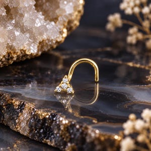 May include: A gold-toned nose stud featuring a triangular cluster of three small, clear, round-cut stones. The jewelry has a curved post design, with the stone setting positioned at the end of the metal wire, reflecting light on its surface.
