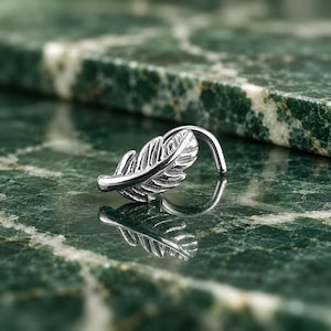 May include: A silver-toned nose stud featuring a detailed feather design with a curved post. The metal surface is polished and reflective, showcasing intricate grooves along the leaf-like structure, creating a distinct and textured appearance.