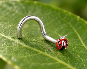 May include: A silver-toned curved nose stud featuring a small ladybug charm at the end. The ladybug is painted with a bright red enamel body, black spots, and a black head, creating a detailed insect design on the metallic jewelry piece.