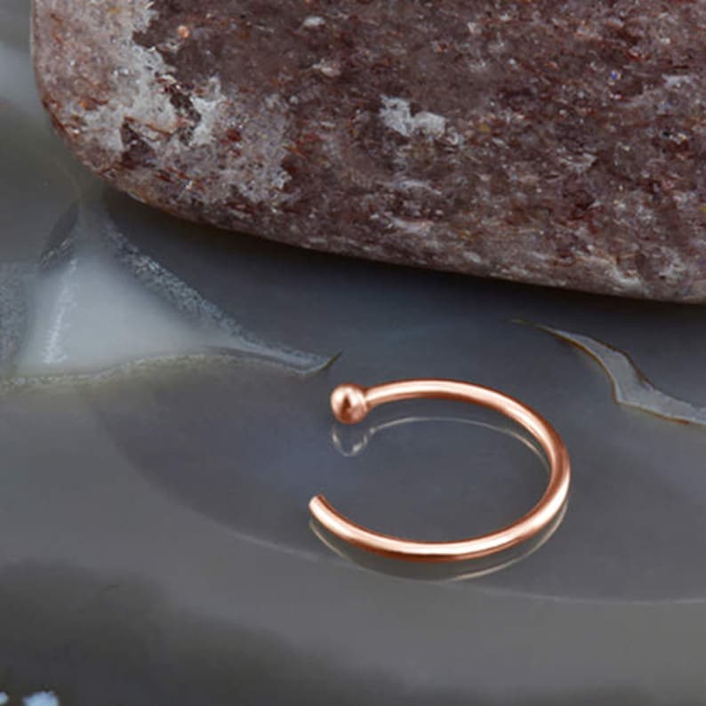 May include: A rose gold nose ring with a small ball at the end. The ring is made of metal and is about 1/4 inch in diameter.