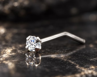 May include: A silver-toned L-shaped nose stud featuring a single round, clear, faceted gemstone held in a four-prong setting. The metal post is bent at a right angle, and the clear stone reflects light with a brilliant, multi-faceted appearance.