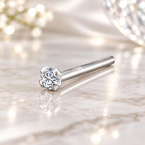 May include: A silver-toned nose stud featuring a single round, clear, faceted stone held in a four-prong setting. The straight post is engraved with the text 925, indicating sterling silver composition, and has a polished, reflective metallic finish.