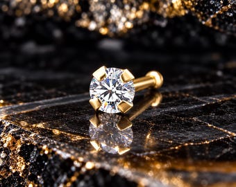 May include: A gold-toned stud earring featuring a single round, clear, faceted gemstone held in a four-prong setting. The post extends backward from the setting, ending in a small, rounded flat back piece, reflecting light on a dark surface.